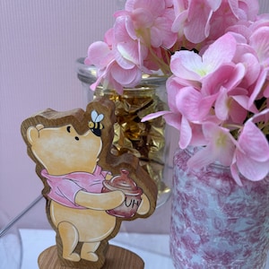May include: A wooden cutout of a yellow bear wearing a pink shirt, holding a brown honey pot labeled with the letters HU, with a small bee perched on its nose, standing on a circular wooden base next to a glass jar filled with pink flowers.