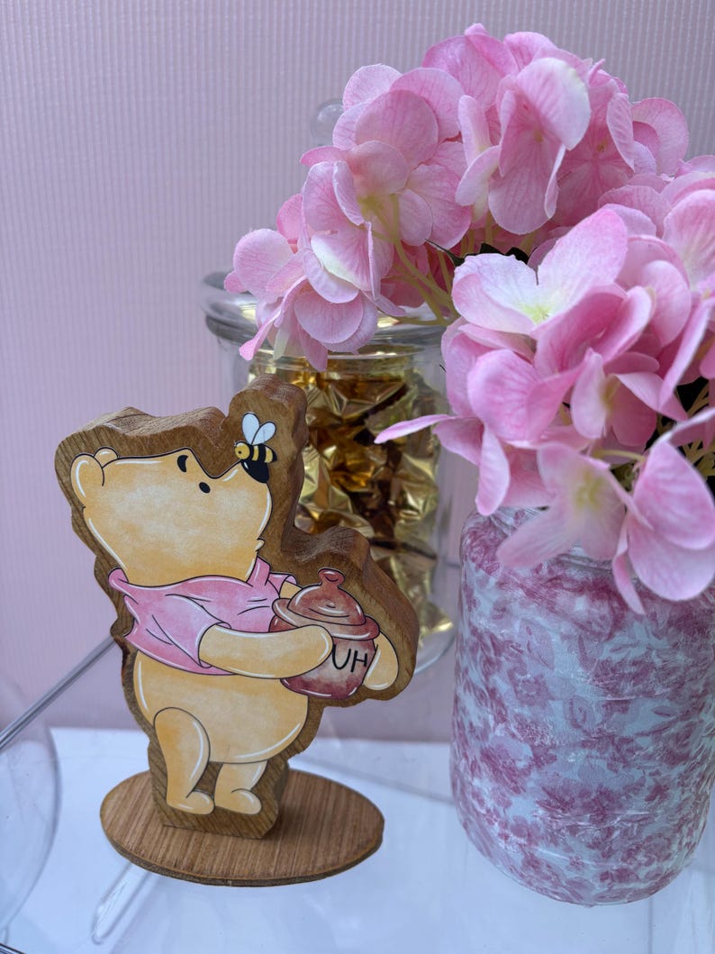 May include: A wooden cutout of a yellow bear wearing a pink shirt, holding a brown honey pot labeled with the letters HU, with a small bee perched on its nose, standing on a circular wooden base next to a glass jar filled with pink flowers.