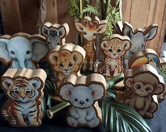 May include: A set of wooden animal figurines painted with realistic details. The animals include a tiger, elephant, zebra, giraffe, monkey, koala, toucan, rhino, and two lion cubs. The figurines are arranged on a dark surface with green leaves and bamboo.