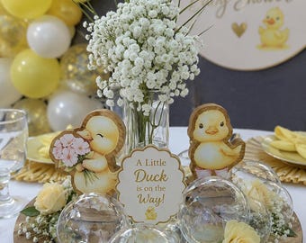 May include: A wooden centerpiece featuring a glass vase with white baby's breath, two duck illustrations, and a central sign reading A Little Duck is on the Way! surrounded by clear plastic spheres and pale yellow roses on a round wooden tray.