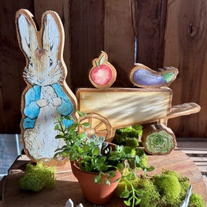 May include: Wooden Peter Rabbit figure with a blue jacket, a small cart, and miniature vegetables. A small terracotta pot with greenery and a white picket fence are also present. The scene is set on a wooden surface.