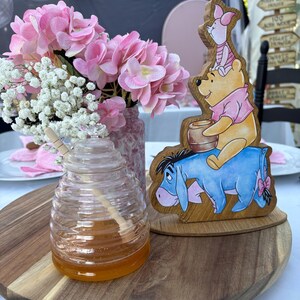 May include: A clear glass beehive-shaped honey jar with a wooden dipper, a wooden cutout featuring Winnie the Pooh, Piglet, and Eeyore, and a vase filled with pink hydrangea and white baby's breath flowers arranged on a round wooden tray.