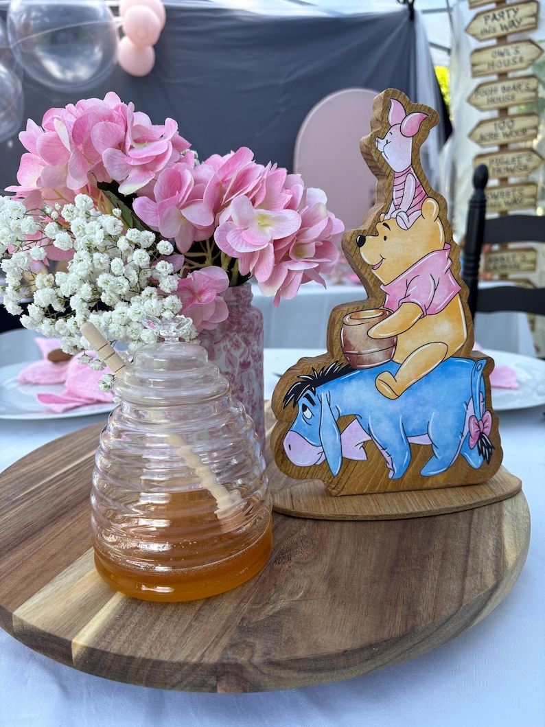 May include: A clear glass beehive-shaped honey jar with a wooden dipper, a wooden cutout featuring Winnie the Pooh, Piglet, and Eeyore, and a vase filled with pink hydrangea and white baby's breath flowers arranged on a round wooden tray.