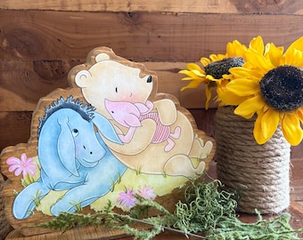 May include: A wooden cutout featuring a light blue donkey and a light tan bear holding a small pink piglet. The characters are illustrated in a soft style, resting on a green grassy patch with small pink flowers. The piece has a natural wood grain finish.