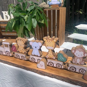 May include: Wooden train set with animal figures, including a tiger, koala, giraffe, and monkey. The train cars are on a wooden base, with heart-shaped accents that read "Be Kind", "Stand Tall", and "Have Fun". The word "BABY" is displayed in white letters.