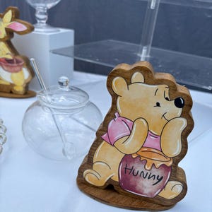 May include: A wooden cutout of a yellow bear wearing a pink shirt, holding a dark red honey pot with the word Hunny written on it. The bear is depicted in a seated position with a light brown wood grain border and a flat base for standing upright.
