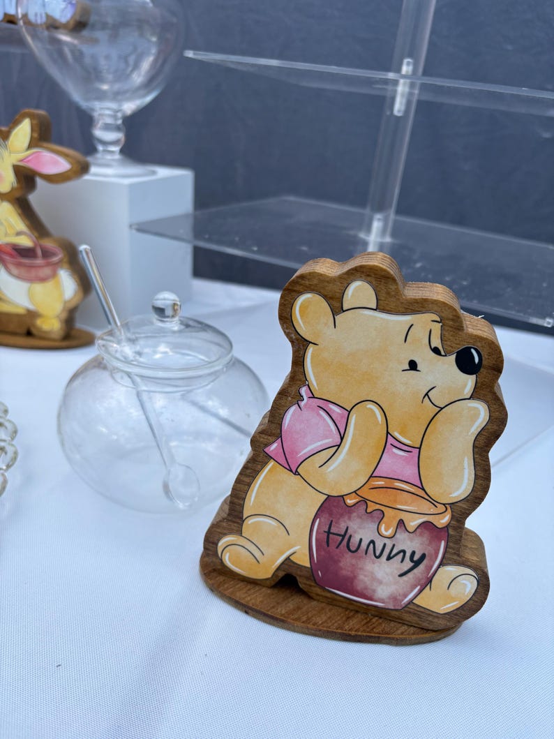 May include: A wooden cutout of a yellow bear wearing a pink shirt, holding a dark red honey pot with the word Hunny written on it. The bear is depicted in a seated position with a light brown wood grain border and a flat base for standing upright.