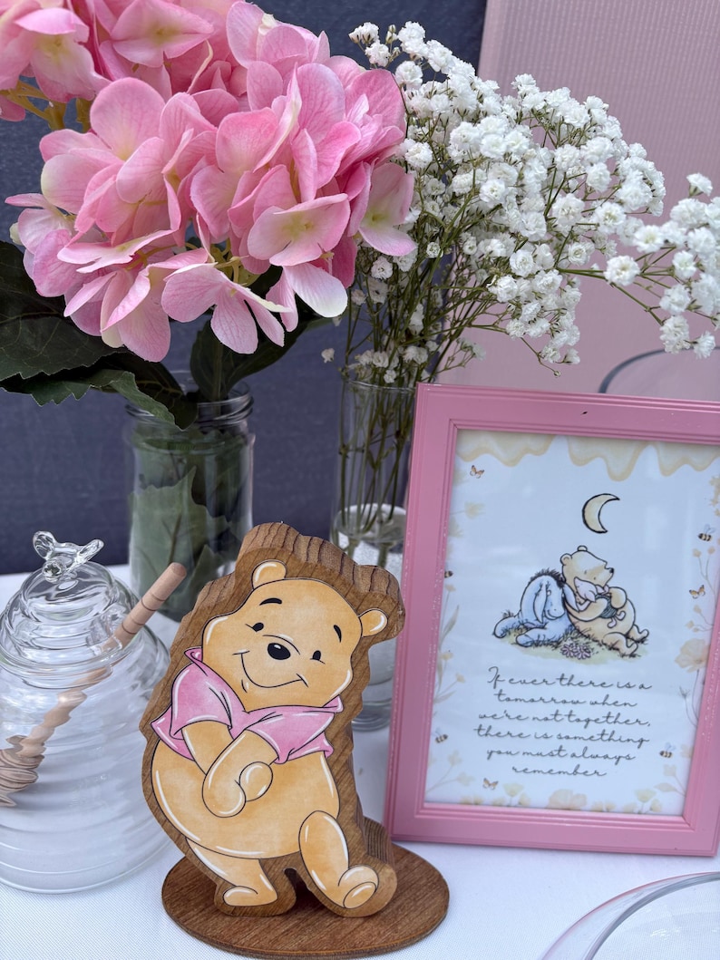 May include: A wooden cutout of Winnie the Pooh wearing a pink shirt, a glass honey jar with a wooden dipper, and a pink-framed print featuring Winnie the Pooh and Piglet with the text: If ever there is a tomorrow when we're not together, there is something you must always remember.