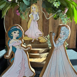 Puede incluir: Tres figuritas de princesas de madera con vestidos rosas. Las princesas son Jasmine, Rapunzel y Elsa. Cada figurita est&aacute; de pie sobre una base de madera.