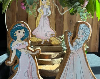 May include: Three wooden princess figurines with pink dresses. The princesses are Jasmine, Rapunzel, and Elsa. Each figurine is standing on a wooden base.