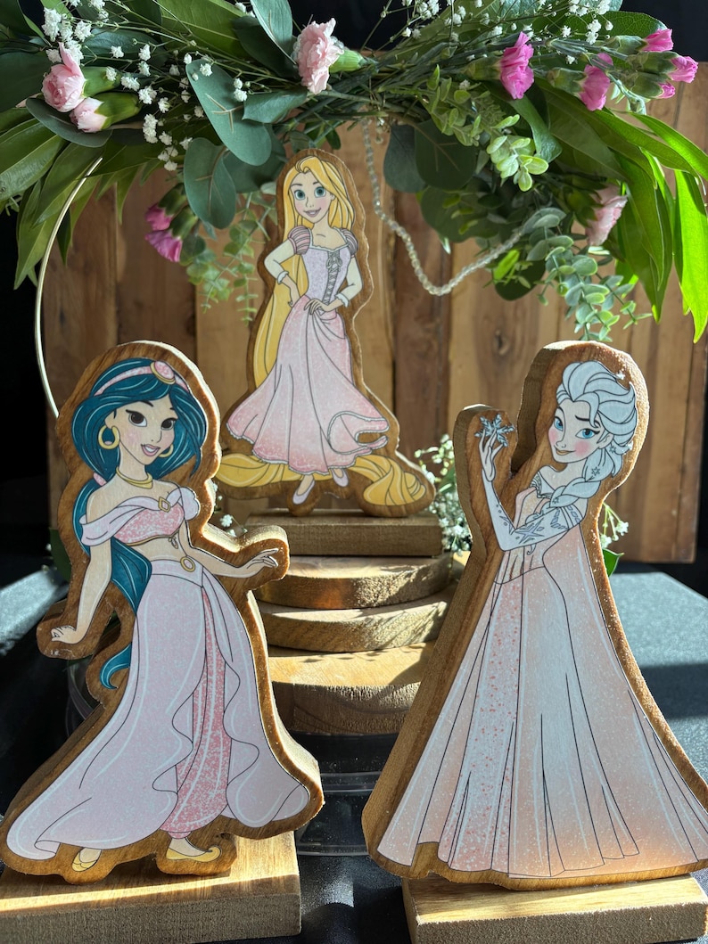 Puede incluir: Tres figuritas de princesas de madera con vestidos rosas. Las princesas son Jasmine, Rapunzel y Elsa. Cada figurita est&aacute; de pie sobre una base de madera.
