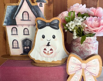 May include: A wooden cutout of a haunted house with pink walls and a black roof, a wooden ghost cutout wearing a black bow and holding a pink pumpkin, and a wooden bow cutout with a pink background and small white ghost patterns.