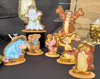 May include: A collection of seven wooden character cutouts featuring Eeyore, Lumpy, Roo, Piglet, Tigger, Rabbit, and Owl. Each figure is painted in vibrant colors and mounted on a small wooden base, showcasing detailed illustrations of the characters.