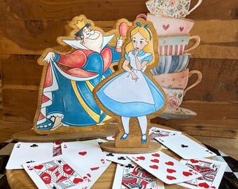 May include: A wooden cutout of the Queen of Hearts and Alice from Alice in Wonderland, accompanied by a stack of illustrated teacups. The figures are arranged on a round wooden base scattered with standard playing cards featuring red and black suits.