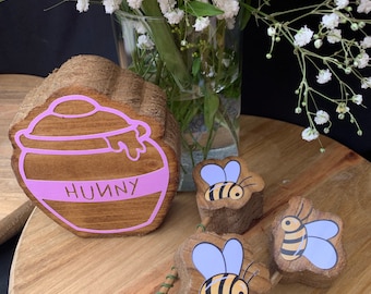 May include: Wooden honey pot with the word "Huny" written on it, surrounded by three wooden bee figurines with purple wings.