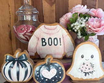 May include: A set of wooden Halloween decorations including a pink sweater cutout with the text BOO!, a pumpkin with black and white stripes and a black bow, a dark pumpkin with a heart pattern, and a ghost holding a bouquet of pink flowers.