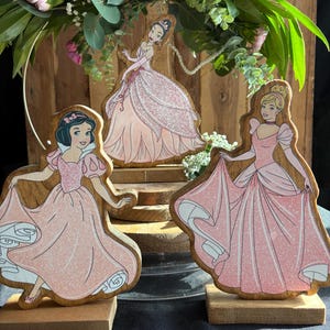 Puede incluir: Tres figuritas de princesas de madera con vestidos rosas y detalles de purpurina. Las princesas son Blancanieves, Cenicienta y Aurora. Las figuritas est&aacute;n sobre bases de madera.