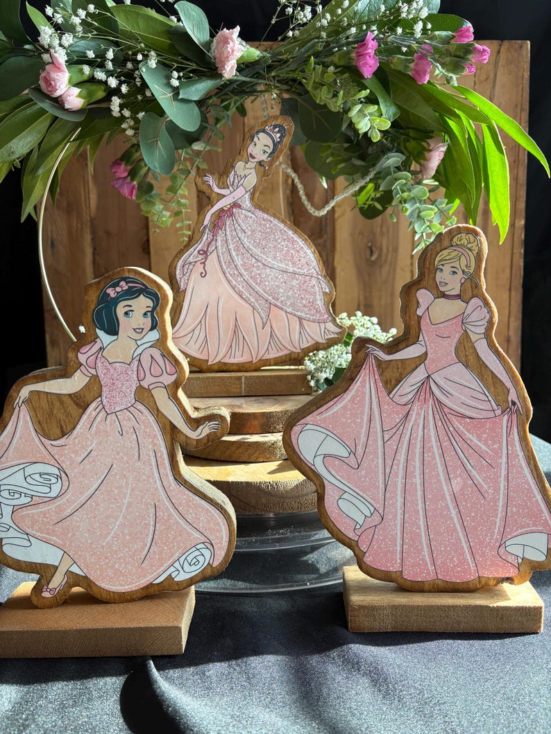 Puede incluir: Tres figuritas de princesas de madera con vestidos rosas y detalles de purpurina. Las princesas son Blancanieves, Cenicienta y Aurora. Las figuritas est&aacute;n sobre bases de madera.