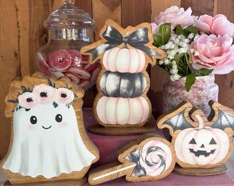 May include: A set of wooden Halloween decorations including a ghost with a floral crown, a stack of three pumpkins with a black bow, a lollipop with a black bow, and a pumpkin with bat wings, all featuring pink, white, and black color schemes.