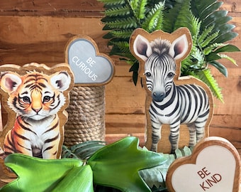 May include: A wooden tiger cutout, a wooden zebra cutout, and two heart-shaped wooden signs. One heart sign features the text BE CURIOUS, and the other heart sign features the text BE KIND. The items are arranged with green artificial foliage.