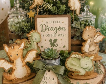 May include: A wooden sign reading A LITTLE DRAGON IS ON HIS WAY! surrounded by several dragon-shaped cutouts in shades of green and tan, a small succulent in a metal pot, and a tag with a green ribbon reading It's a Boy! on a wood slice.