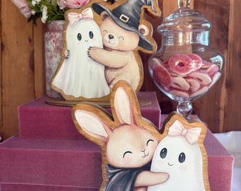 May include: Two wooden cutout decorations featuring illustrated characters. One shows a light brown bear wearing a black witch hat hugging a white ghost with a pink bow. The other shows a light brown rabbit wearing a black cape hugging a white ghost with a pink bow.