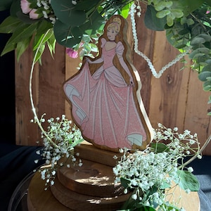 Puede incluir: Una figura de madera de Cenicienta con un vestido rosa y un collar de perlas blancas, rodeada de flores blancas y rosas y vegetaci&oacute;n. La figura est&aacute; sobre un soporte de madera de varios niveles.