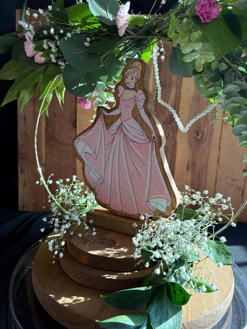 Puede incluir: Una figura de madera de Cenicienta con un vestido rosa y un collar de perlas blancas, rodeada de flores blancas y rosas y vegetaci&oacute;n. La figura est&aacute; sobre un soporte de madera de varios niveles.