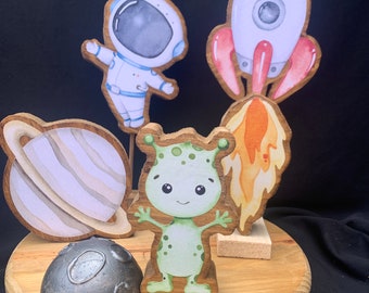 May include: Wooden space-themed decorations including an astronaut, rocket, planet Saturn, a green alien, and a gray moon. The figures are painted with watercolor details and set on a wooden base, perfect for a child's room or space-themed party.