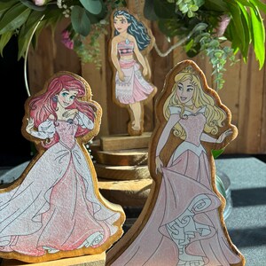 Puede incluir: Tres figuritas de princesas de madera, cada una con una princesa diferente de Disney. Las princesas est&aacute;n de pie sobre bases de madera. Las princesas son Moana, Ariel y Aurora. Las princesas llevan todas vestidos rosas.