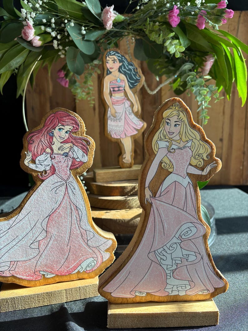 Puede incluir: Tres figuritas de princesas de madera, cada una con una princesa diferente de Disney. Las princesas est&aacute;n de pie sobre bases de madera. Las princesas son Moana, Ariel y Aurora. Las princesas llevan todas vestidos rosas.