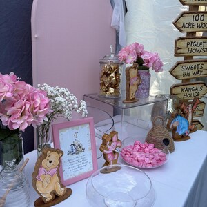 May include: A Winnie the Pooh themed party display featuring a pink arched backdrop, a wooden signpost with directional arrows, pink hydrangea bouquets, glass jars filled with candies, and wooden character cutouts of Pooh, Piglet, and Eeyore.