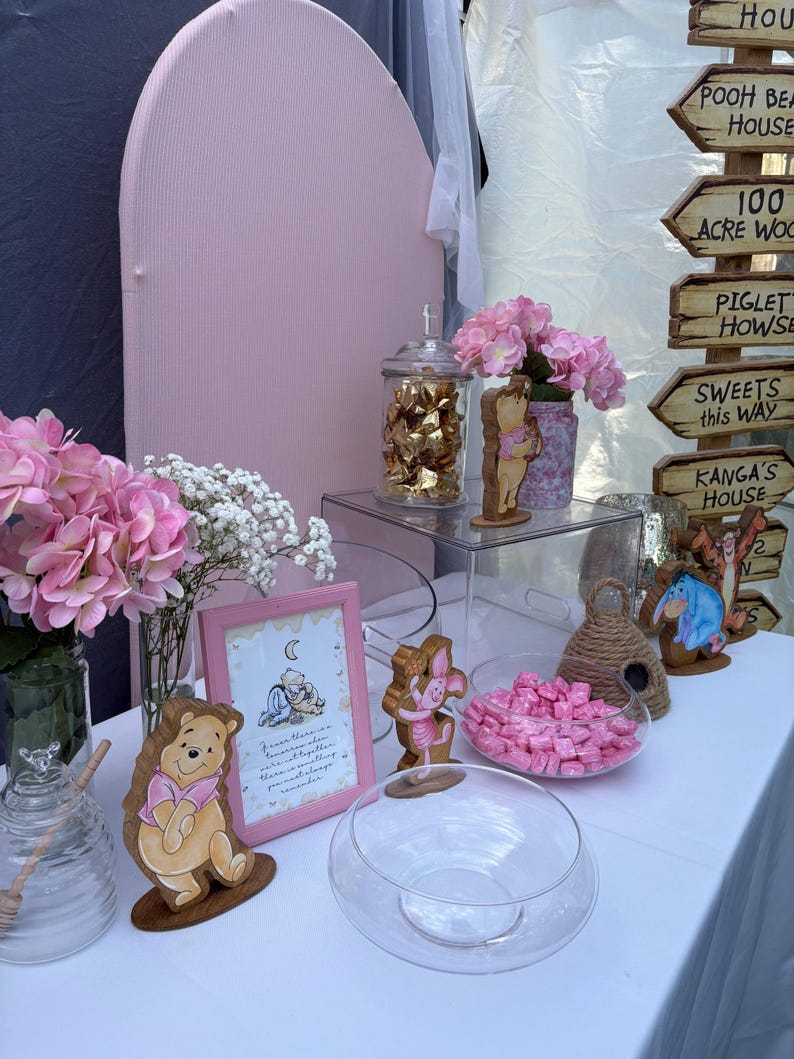 May include: A Winnie the Pooh themed party display featuring a pink arched backdrop, a wooden signpost with directional arrows, pink hydrangea bouquets, glass jars filled with candies, and wooden character cutouts of Pooh, Piglet, and Eeyore.