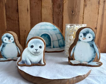 May include: Wooden figurines of a penguin, a seal, and an igloo. The penguin and seal are painted with realistic details, and the igloo is painted with blue and white stripes. The figurines are arranged on a wooden base.