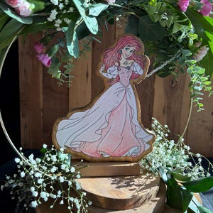 Puede incluir: Una figurita de madera de Ariel de La Sirenita, con un vestido rosa y de pie sobre una base de madera de varios niveles. La figurita est&aacute; rodeada de vegetaci&oacute;n y flores blancas.