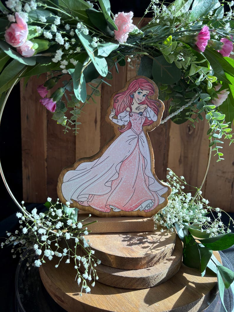 Puede incluir: Una figurita de madera de Ariel de La Sirenita, con un vestido rosa y de pie sobre una base de madera de varios niveles. La figurita est&aacute; rodeada de vegetaci&oacute;n y flores blancas.