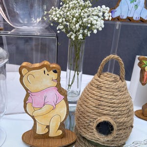 May include: A wooden cutout of a yellow bear wearing a pink shirt standing on a circular base, positioned next to a dome-shaped beehive made of thick brown jute rope with a small circular opening at the front, and a clear glass vase with white flowers.