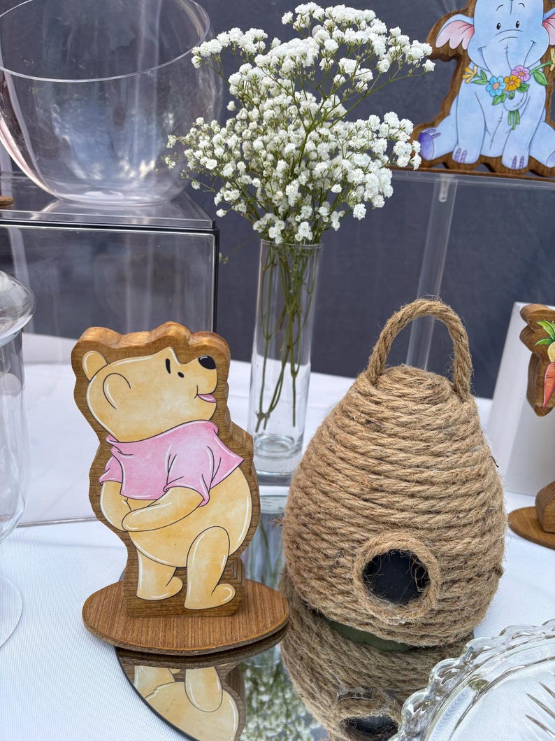 May include: A wooden cutout of a yellow bear wearing a pink shirt standing on a circular base, positioned next to a dome-shaped beehive made of thick brown jute rope with a small circular opening at the front, and a clear glass vase with white flowers.