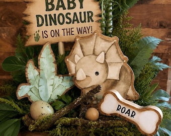 May include: A wooden centrepiece featuring a sign that reads BABY DINOSAUR IS ON THE WAY!, a triceratops cutout, a bone-shaped sign with the text ROAR, a plant cutout, two small eggs, and green foliage arranged on a round wood slice base.