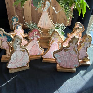 Puede incluir: Una colecci&oacute;n de figuritas de princesas de madera, cada una pintada con un personaje de princesa diferente con un vestido rosa. Las figuritas est&aacute;n dispuestas sobre una base de madera y est&aacute;n rodeadas de vegetaci&oacute;n.