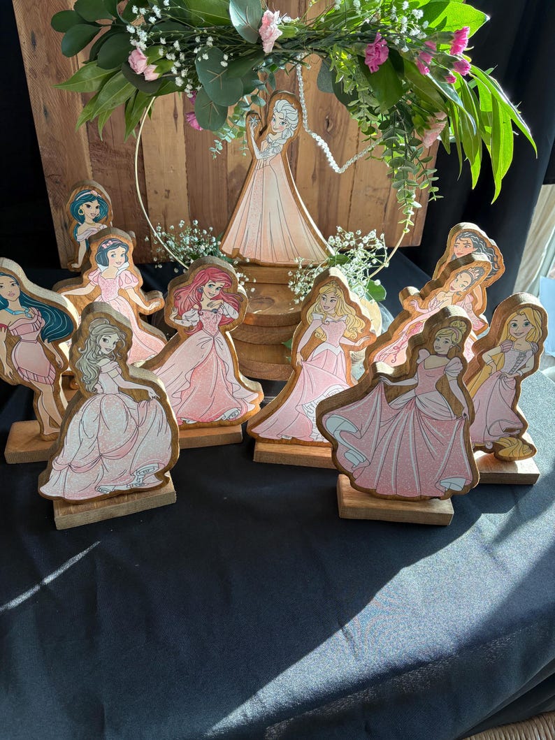 Puede incluir: Una colecci&oacute;n de figuritas de princesas de madera, cada una pintada con un personaje de princesa diferente con un vestido rosa. Las figuritas est&aacute;n dispuestas sobre una base de madera y est&aacute;n rodeadas de vegetaci&oacute;n.