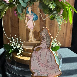 Puede incluir: Dos figuritas de princesas de madera, una con un vestido rosa y la otra con un vestido azul, se exhiben en un soporte de madera de varios niveles. El soporte est&aacute; decorado con una corona de flores.