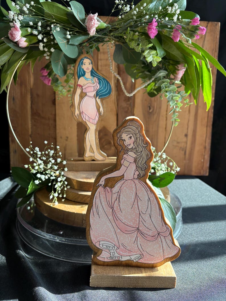 Puede incluir: Dos figuritas de princesas de madera, una con un vestido rosa y la otra con un vestido azul, se exhiben en un soporte de madera de varios niveles. El soporte est&aacute; decorado con una corona de flores.