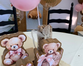 May include: A wooden centerpiece featuring two teddy bears, one sitting on a book and the other in a basket. The centerpiece also includes a wooden gift box with a pink bow and two wooden rings. The centerpiece is decorated with a clear acrylic sign that reads "We can bearly wait".