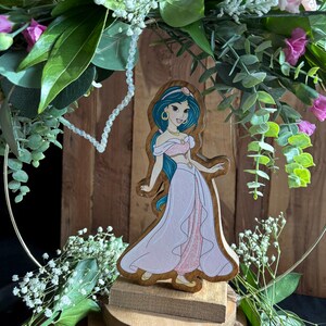 Puede incluir: Una figurita de madera de la princesa Jasmine de Aladdin de Disney. Lleva un atuendo azul y rosa y tiene el pelo largo y oscuro. La figurita est&aacute; sobre una base de madera y est&aacute; rodeada de vegetaci&oacute;n.