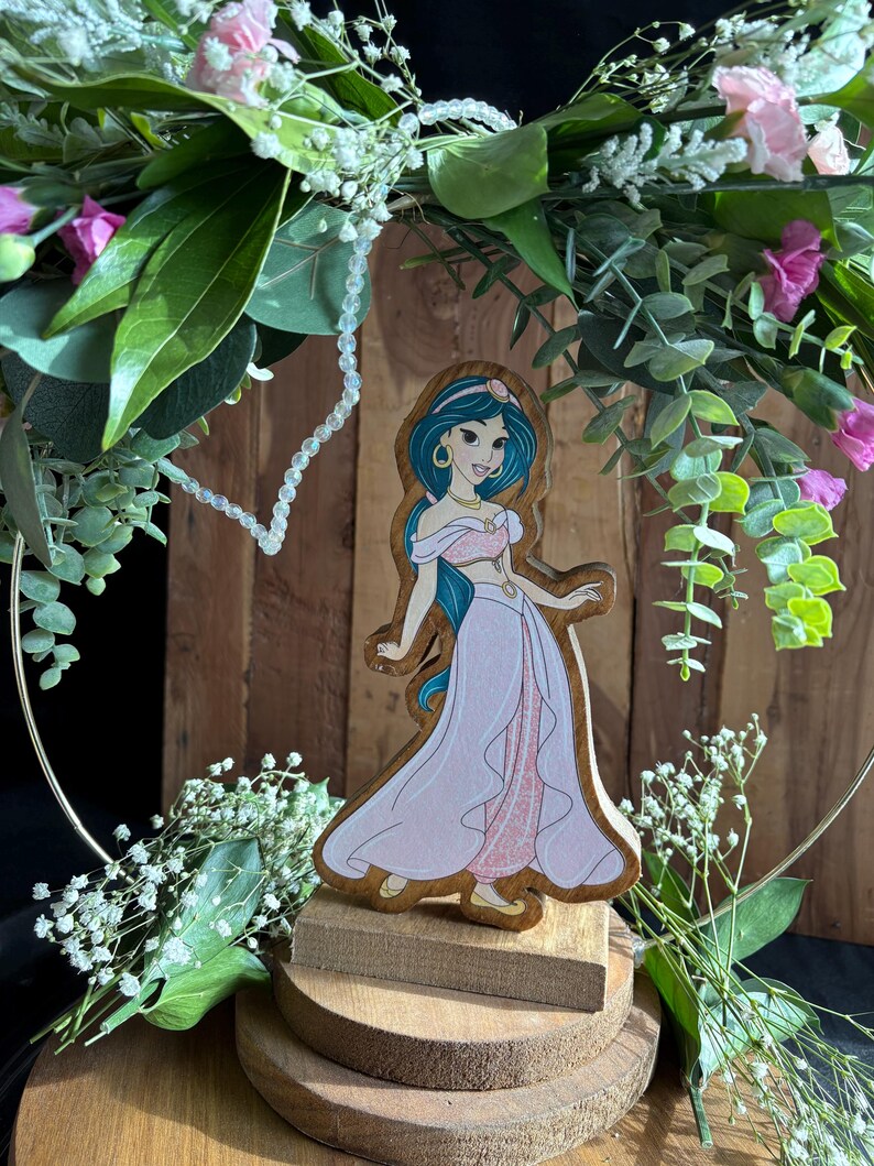 Puede incluir: Una figurita de madera de la princesa Jasmine de Aladdin de Disney. Lleva un atuendo azul y rosa y tiene el pelo largo y oscuro. La figurita est&aacute; sobre una base de madera y est&aacute; rodeada de vegetaci&oacute;n.