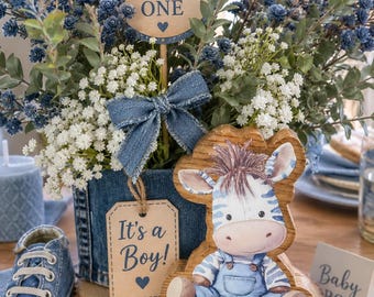 May include: A decorative arrangement featuring a blue denim-wrapped container, blue and white flowers, and a round sign reading WELCOME little ONE. A wooden cutout of a zebra in overalls, a denim bow, and a tag reading It's a Boy! accompany a blue sneaker.