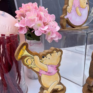 May include: Two wooden Winnie the Pooh cutouts featuring the character in a pink shirt, one playing a yellow trumpet and the other resting on a pink balloon. A glass vase filled with red licorice and a bouquet of light pink hydrangea flowers.