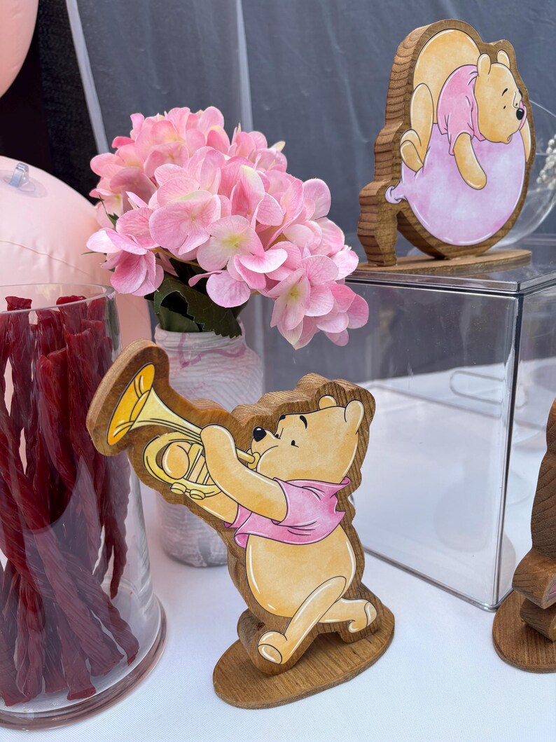 May include: Two wooden Winnie the Pooh cutouts featuring the character in a pink shirt, one playing a yellow trumpet and the other resting on a pink balloon. A glass vase filled with red licorice and a bouquet of light pink hydrangea flowers.
