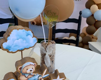 May include: A wooden airplane centerpiece with a teddy bear pilot, blue and white clouds, and a star. The centerpiece is surrounded by brown and beige balloons with the text "We can barely wait" on one of the balloons.